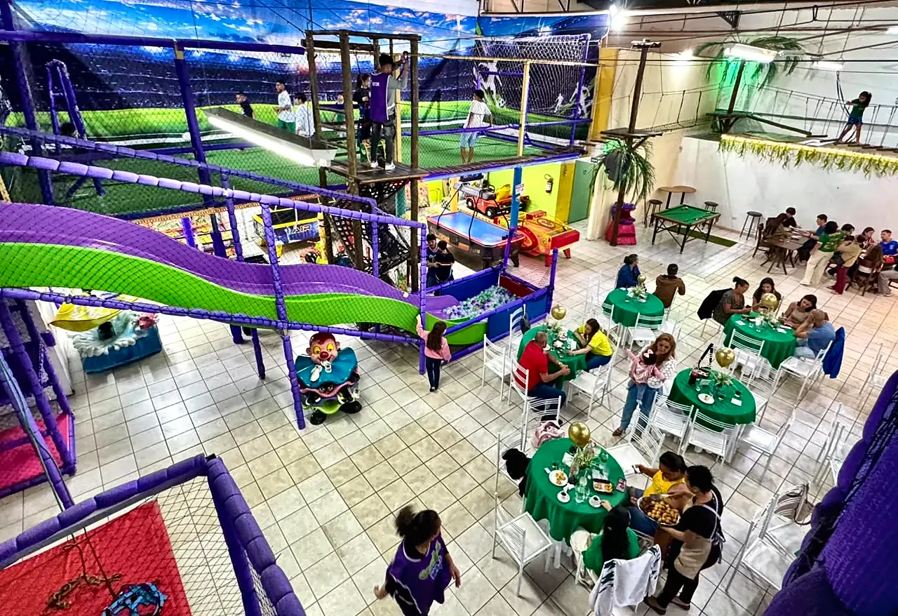 Vista do alto do salão durante uma festa: convidados à mesa, tobogã, arvorismo e campo de futebol ao fundo